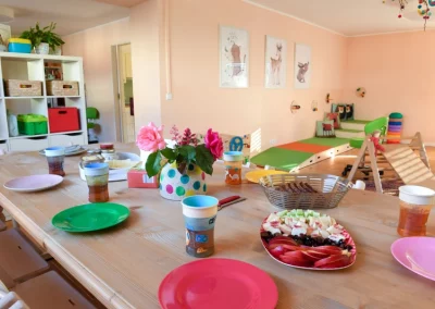 Spielzimmer mit gedecktem Frühstückstisch mit bunten Tellern, Obst und Brot. Im Hintergrund bunte Möbel zum Spielen, Bilder mit lustigen Motiven hängen an der Wand.