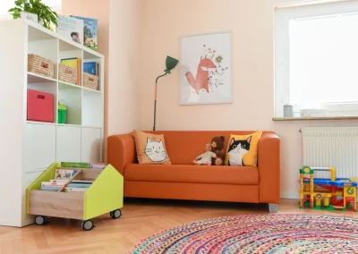 Teil des Spielzimmers mit Sofa, Büchern und Spielzeug, ein bunter Teppich im Vordergrund.