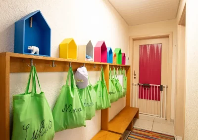 Flur im Eingang mit bunter Garderobe, die Tür ist mit einem Schutzgitter gesichert, für jedes Kind hängt eine Tasche mit Namen am Garderobenhaken.