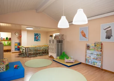 Blick auf den Eingang im Hintergrund, dann der Esstisch, rechts steht ein Kachelofen, dvor eine Matratze mit Spielzeug und ein Regal mit Bilderbüchern. Heller Boden mit runden Teppichen.