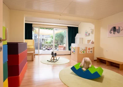 Ein weiteres Spielzimmer mit bunten Spiel-Matratzen links und einer Bank und Spielzeug an den Wänden rechts. Im hinteren Teil der Zugang zur Terrasse.